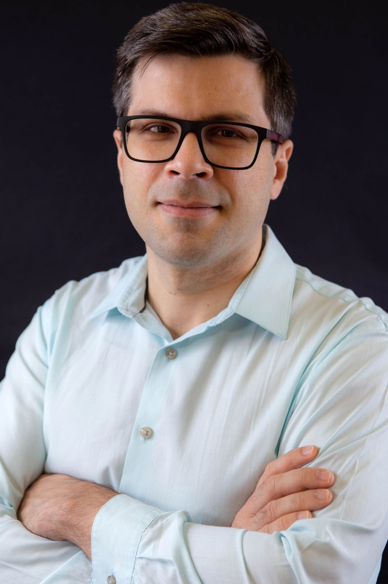 Person wearing a light blue dress shirt with arms crossed against a dark background.